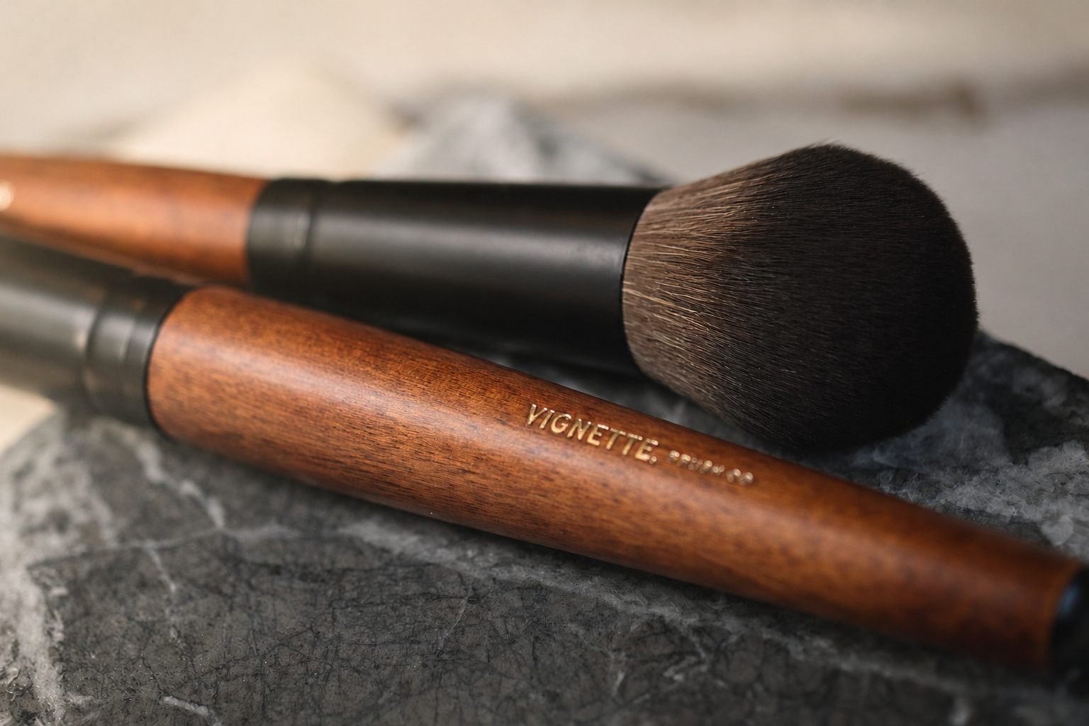 Two makeup brushes with wooden handles and black ferrules on a textured surface.
